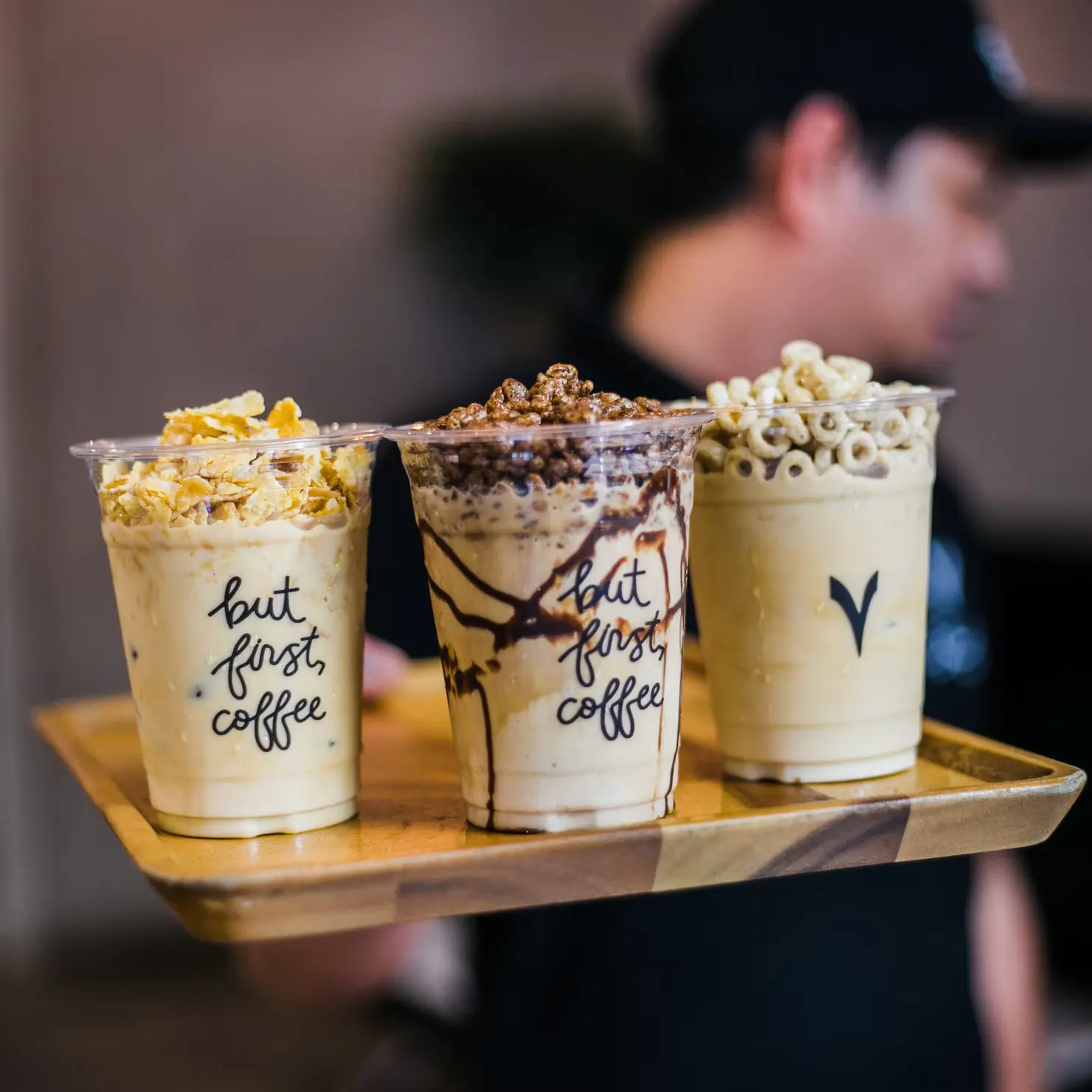 Out of focus man with long hair holding three milkshakes topped with cereal up to the camera.